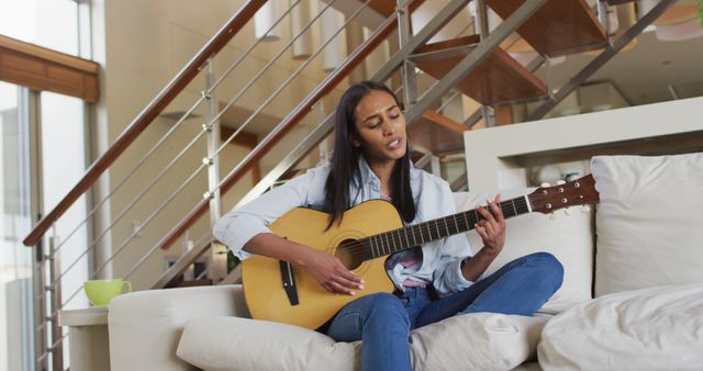 Young woman enjoys playing guitar while sitting comfortably on a couch in a stylish, modern living room with an open staircase in the background. Perfect for depicting leisure time, home comfort, musical hobbies, and modern interior design. Useful for lifestyle blogs, home decor websites, and advertisements related to music or relaxation products.