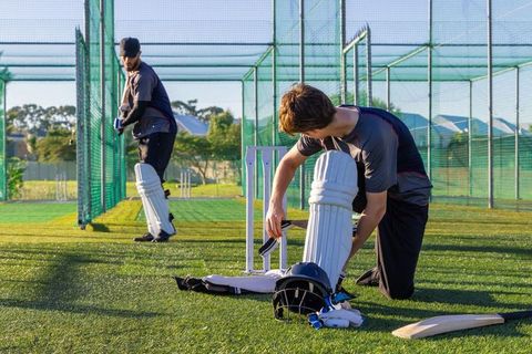 Cricket teammates practicing batting in outdoor nets with pads helmet bat and stumps