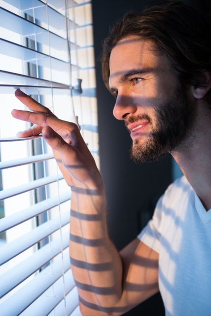 Man peeking through window blinds with morning sunlight casting shadows on his face. Ideal for concepts of curiosity, morning routines, home life, and relaxation.