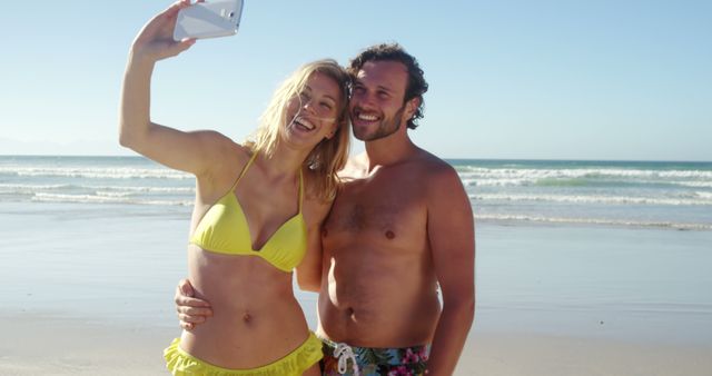 Young couple standing on sandy beach, posing for cheerful selfie with ocean waves in background. Use for vacation promotions, summer travel blogs, lifestyle advertisements, or social media campaigns highlighting carefree fun and memorable moments at the beach.