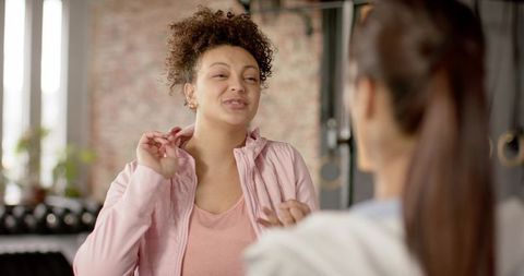 Women talking after workout in sunlit gym wearing pink jacket sharing fitness motivation