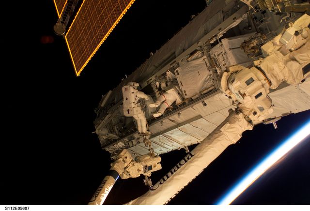 Astronauts are conducting maintenance on the International Space Station during a spacewalk. The newly installed Starboard One (S1) Truss is visible, with sunlight glinting at Earth's horizon. This image can be used in educational materials about space exploration, scientific articles on astronauts and space missions, and promotional content for space-related activities and organizations.