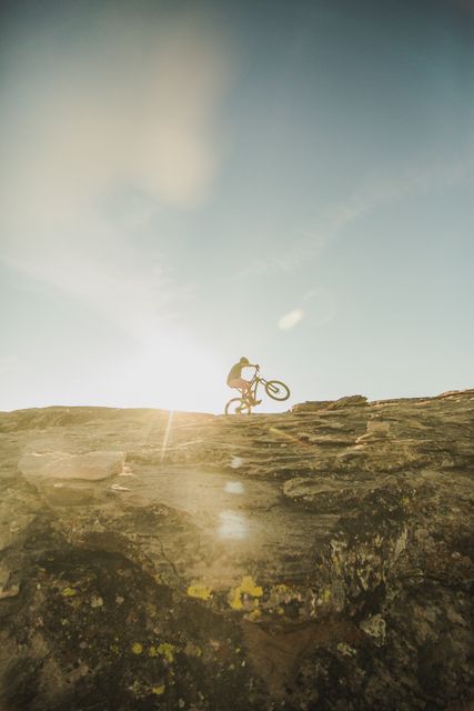 Man cycling on rocky terrain with sun flare shining in the background. Ideal for promoting outdoor adventures, extreme sports, fitness activities, and nature explorations. Can be used in magazines, brochures, and websites focused on sports or outdoor activities.