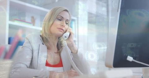Businesswoman speaking on corded telephone at desk while working on desktop computer