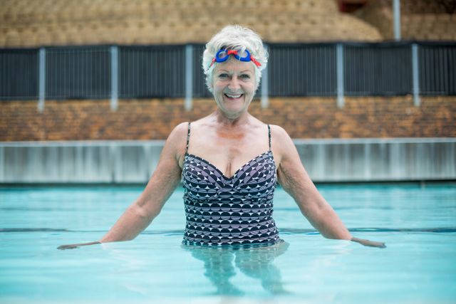 This image depicts a cheerful senior woman enjoying a swim in a pool. She is smiling and appears to be in good spirits, promoting an active and healthy lifestyle for elderly individuals. This image can be used for health and wellness campaigns, fitness programs for seniors, advertisements for swimming pools or swimwear, and articles on active aging and senior fitness.