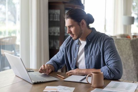 Focused remote worker using laptop and notebook at sunlit dining table