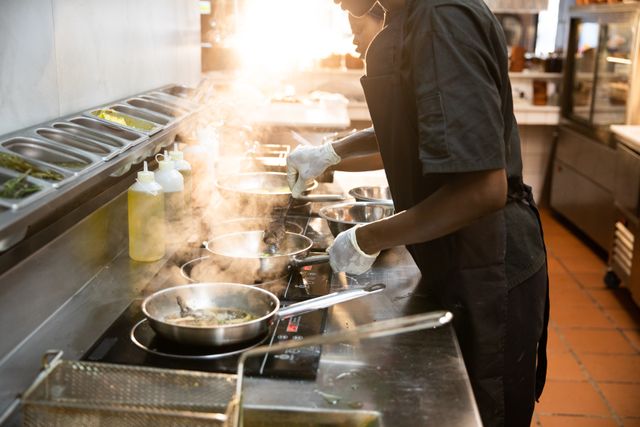 African American male and female chefs are cooking in a busy restaurant kitchen, frying food on multiple frying pans with steam rising. This image is perfect for illustrating teamwork, culinary arts, and professional kitchen environments. It can be used in articles about restaurant operations, culinary schools, chef training, and food preparation.