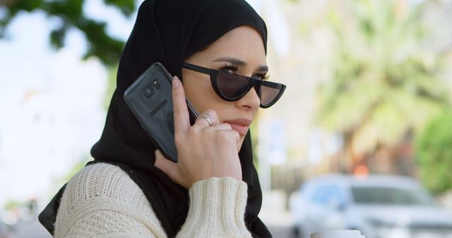 Confident woman wearing hijab and sunglasses is talking on her smartphone while standing outdoors and holding a coffee cup. Great for themes related to modern Muslim women, business, urban life, and technology. This image may be used in articles, advertisements, or online content focusing on lifestyle, communication, and cultural diversity.