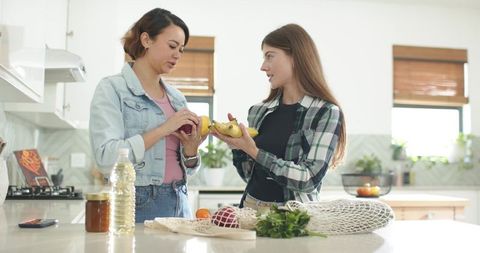 Two women comparing bananas at bright modern kitchen island while unpacking groceries