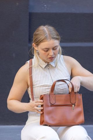Young woman reaching into tan leather handbag while sitting on urban concrete ledge