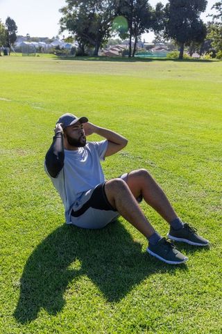 African american man doing crunches in sunny park wearing cap compression sleeve sneakers