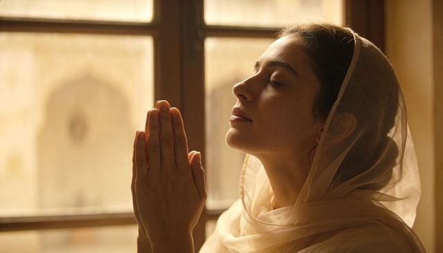 Young woman praying with sheer headscarf by wooden window in warm golden light