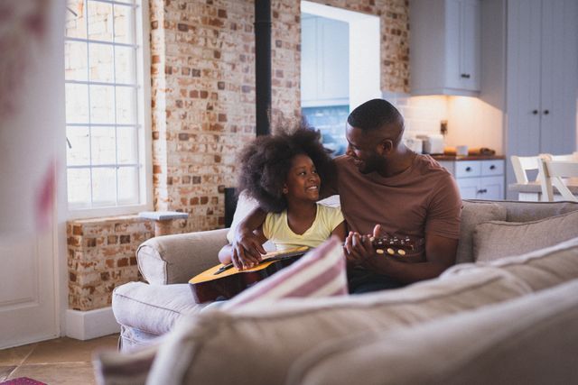 Happy african american girl and her father sitting on couch playing guitar together in living room. staying at home in isolation during quarantine lockdown.