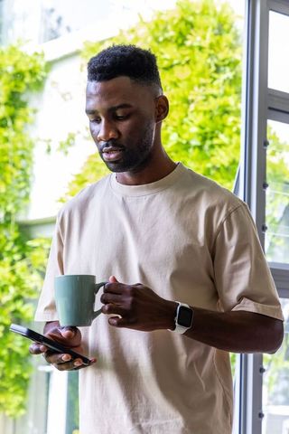 Standing at open window holding mug and phone, african man wearing smartwatch in sunlit home