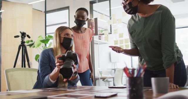 Depicts a creative team collaborating in an office environment while adhering to pandemic guidelines by wearing masks. A person is holding a camera, suggesting a focus on photography or visual content creation. Use this for articles or marketing materials related to workplace safety during the pandemic, modern office environments, or teamwork in creative industries.