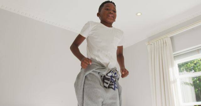 Happy boy jumping on bed in light-filled room dressed in casual clothing. Captures playful moment full of energy and joy. Great for lifestyle, childhood memories, home fun activities, health and fitness campaigns.