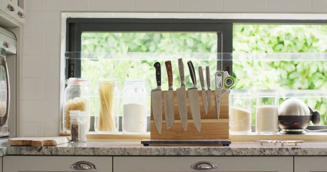High-quality stock image showing a well-organized modern kitchen countertop. Prominent knife set with various utensils placed on a sleek marble counter. Storage jars containing pasta, flour, rice, and grains line against a large window, adding a touch of elegance. Ideal for articles and content related to cooking, home organization, kitchen design, and culinary tools.