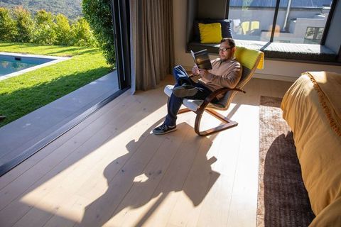 Mature asian man reading paperback in sunlit bedroom chair overlooking pool patio, relaxing