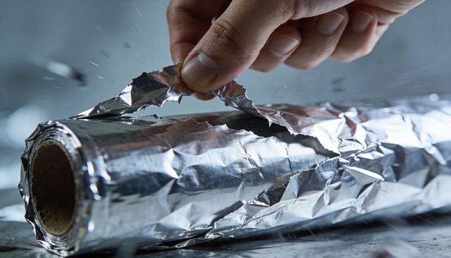 Pinching hand tearing aluminum foil roll on crumpled reflective surface close-up kitchen