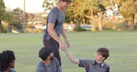 Young male teammates helping each other after outdoor practice on grassy field at sunset