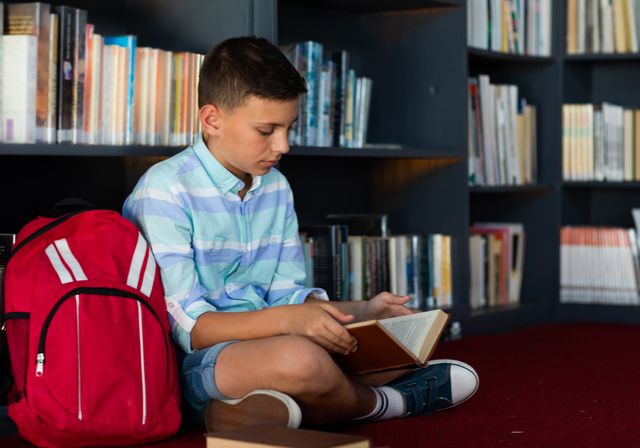 Caucasian schoolboy reading book, sitting on floor with bag in elementary school library. Education, childhood and learning concept.