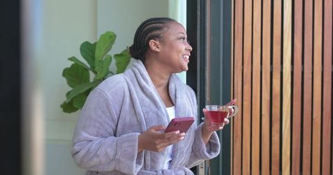 African american woman smiling in robe on balcony holding smartphone and cup of tea