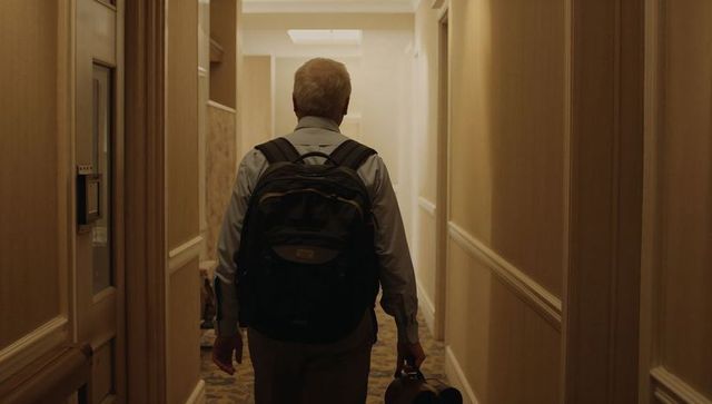 Solo senior traveler walking down hotel corridor carrying backpack and carry-on luggage