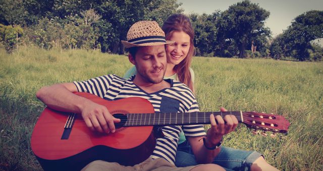A young Caucasian couple enjoys a relaxing moment in a grassy field, with the man playing guitar and the woman smiling behind him, with copy space. Their shared enjoyment of music creates a romantic and serene atmosphere.