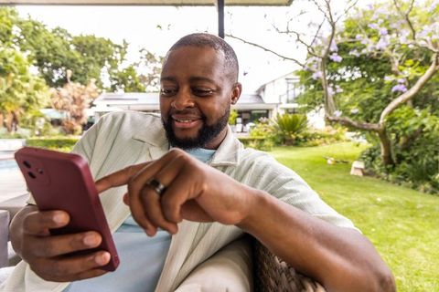 African american man relaxing on patio tapping red smartphone smiling and lounging outside