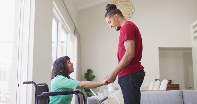Happy biracial woman in wheelchair and smiling male partner holding hands and talking in living room. wellbeing and domestic lifestyle with physical disability.