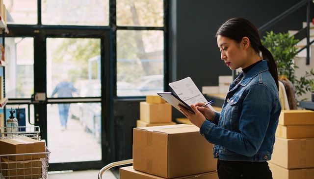 Woman checking inventory on clipboard while organizing boxes at fulfillment packing station