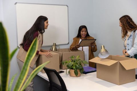 Female coworkers unpacking boxes during office move celebrating new workspace reveal