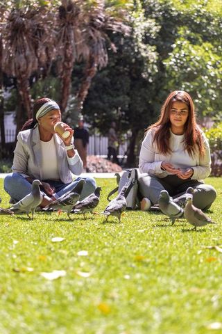 Two women feeding pigeons in sunny urban park cross-legged with takeaway cup and backpacks