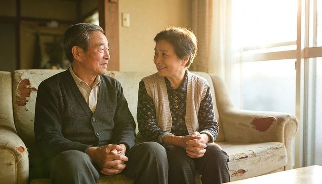 Senior asian couple sitting and smiling on worn sofa in sunlit cozy living room