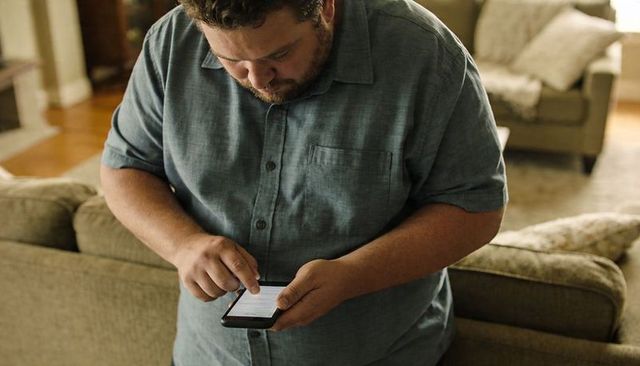 Man tapping smartphone while standing behind sofa in cozy living room, home technology