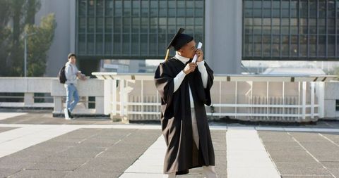 Graduate celebrating on campus plaza holding diploma and smartphone in regalia