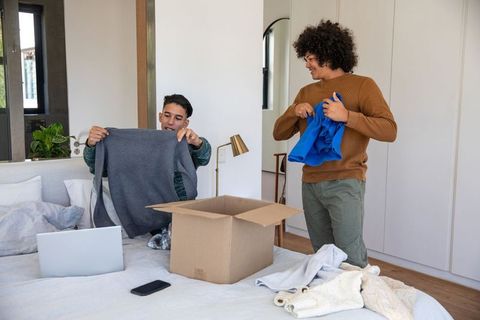 Diverse male friends unpacking clothes from cardboard box on double bed