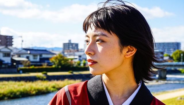 Sunlit woman wearing red kimono gazing toward riverside cityscape and bridge
