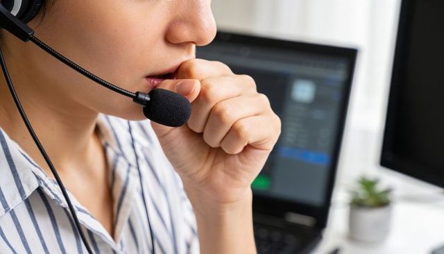Customer support agent pausing on call wearing headset at home workstation with laptop