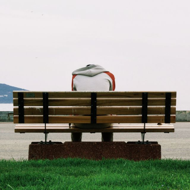 A person is sitting on a wooden bench facing a waterfront, head bent down suggesting contemplation or solitude. This photo could be used in themes of loneliness, introspection, mental health, relaxation in nature, and being in a tranquil coastal environment.