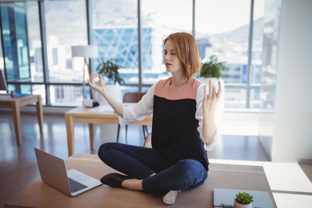 Executive woman practicing mindfulness while sitting cross-legged on office desk. Promotes relaxation and stress relief in work environment. Useful for themes of work-life balance, corporate wellness, and self-care.
