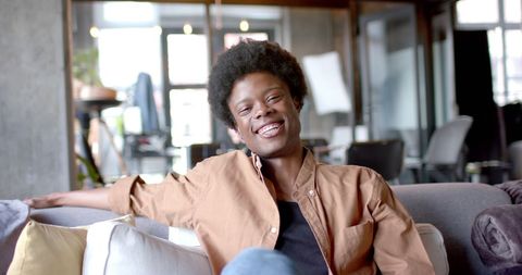 Person with a cheerful expression is sitting on a sofa in a modern living room. Use this image for lifestyle blogs, home comfort themes, or articles focused on personal relaxation and happiness.