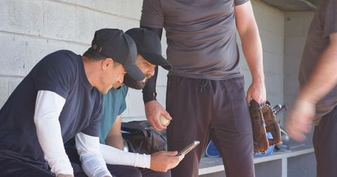Baseball teammates in sunlit dugout viewing smartphone and sharing glove during game break