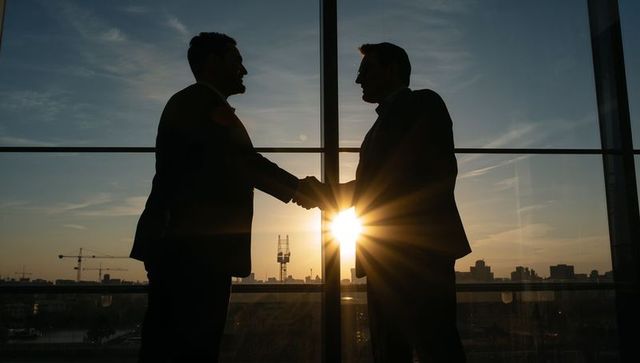 Silhouetted businessmen shaking hands at sunset through full-height office window with city skyline