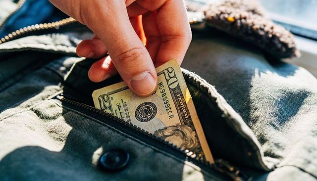 Hand placing folded us twenty dollar bill into olive green jacket pocket by window