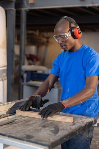 Woodworker wearing goggles and earmuffs cutting wooden plank on table saw in carpentry shop