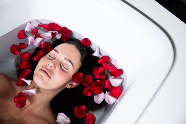 A young Caucasian brunette woman lying in a bathtub with red and pink rose petals floating around her. She has her eyes closed, giving a sense of calmness and relaxation. Perfect for illustrating concepts related to spa treatments, self-care, wellness routines, and peaceful moments. Ideal for use in beauty, health, and lifestyle blogs, as well as promotional material for spas and wellness services.