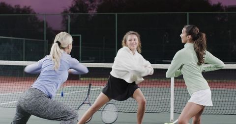 Diverse women teammates stretching on tennis court at dusk warming up together