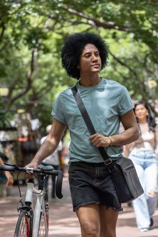 African american man walking bike through leafy urban plaza in teal tee with crossbody bag