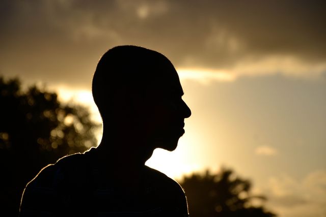 Silhouette of a man with closely cropped hair standing outdoors at sunset. The sky is cloudy, and the silhouette of the man is highlighted by the warm, setting sun. Trees are visible in the background, adding to the peaceful scene. This visual can be used for themes related to introspection, solitude, nature, and sunset.
